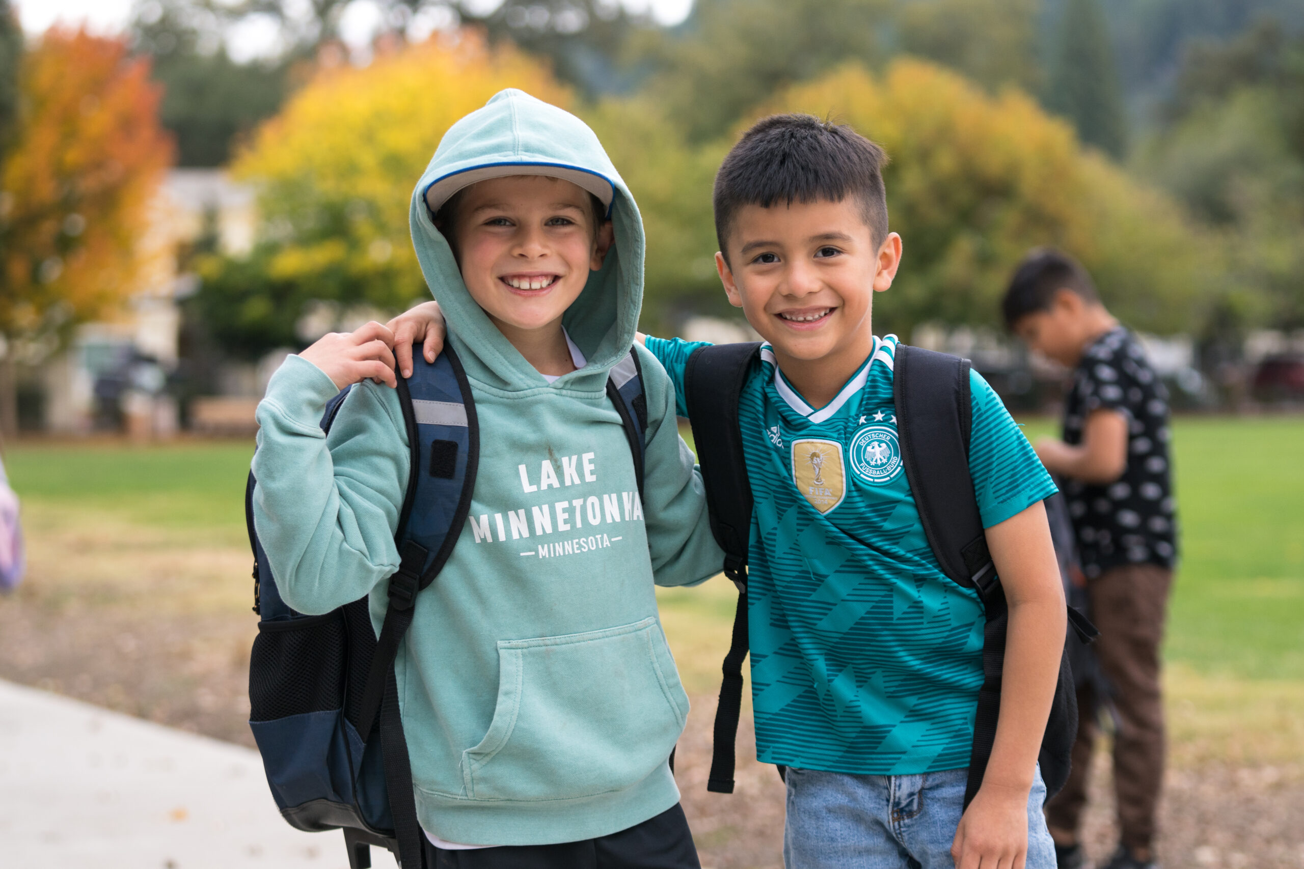 Two smiling kids with backpacks hugging outside.