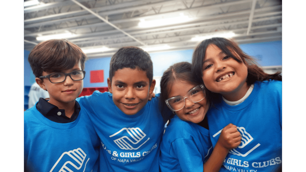 Four smiling children in blue shirts indoors.